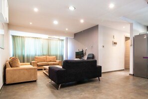 En moderne stue med sofa, lænestol og et lille bord. Der er et køkkenområde med et køleskab og en mikroovn.