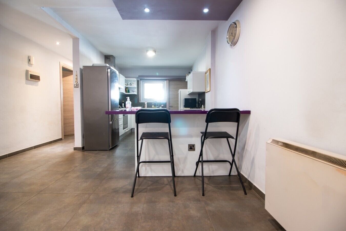 Una cocina moderna con una isla central, dos taburetes de bar, un refrigerador y un reloj en la pared.