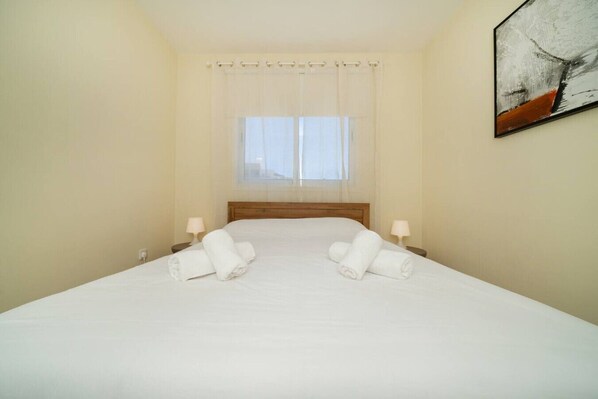 A hotel room with a large bed, two bedside lamps, a window with sheer curtains, and a painting on the wall.