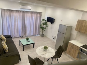 Un salon moderne comprenant un canapé, une table basse, un téléviseur et un coin cuisine avec des placards et un réfrigérateur.