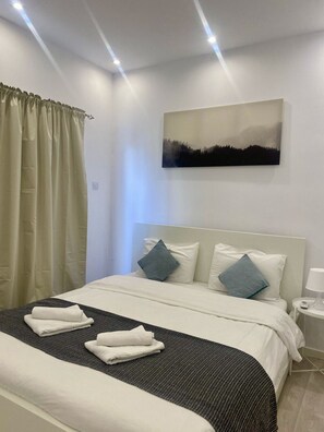 Een net opgemaakt bed met witte lakens en twee blauwe kussens, een boslandschap op de muur en een witte nachtkastje met een lamp.