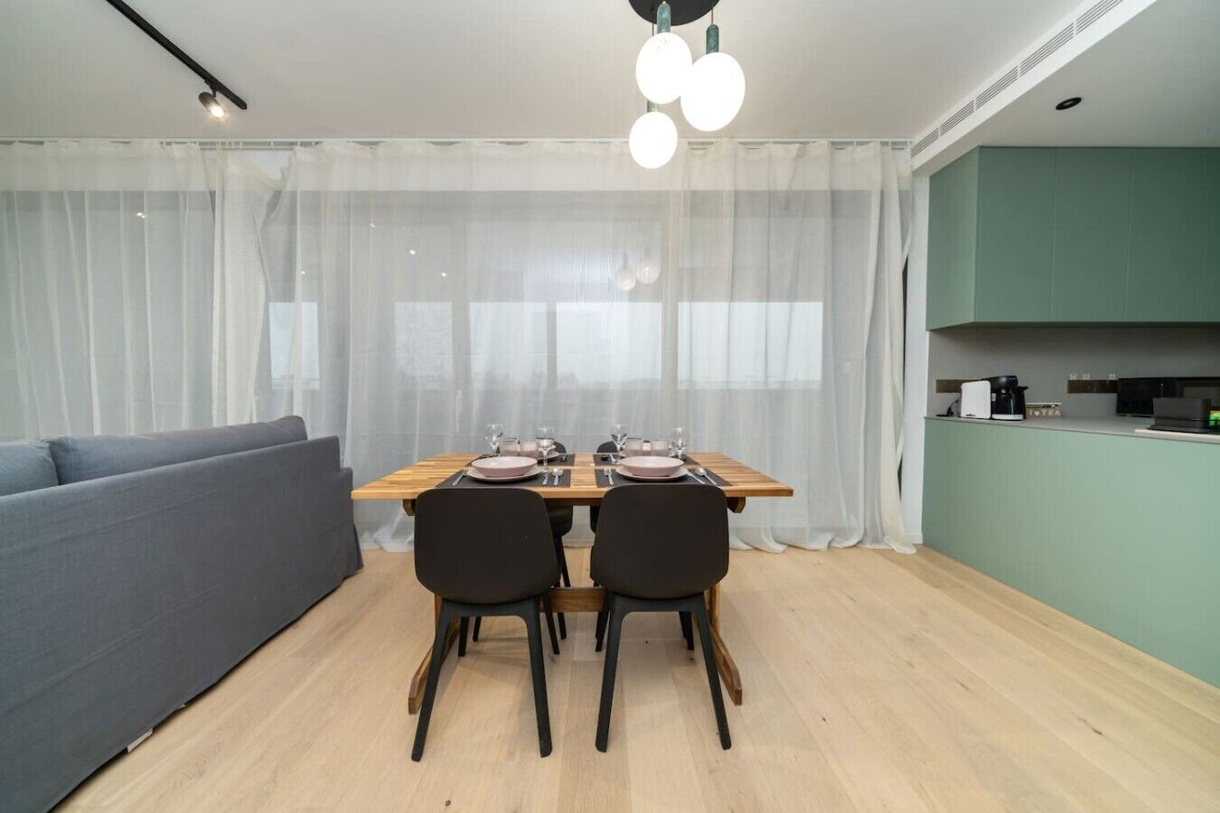 Un espace repas moderne avec une table en bois dressée pour deux personnes, un canapé gris et une cuisine en arrière-plan.