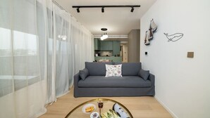 A living room with a grey sofa, a coffee table with snacks, sheer curtains, and a kitchen in the background. - Amazing new 2 bedroom flat near casino beach mall (Limassol)