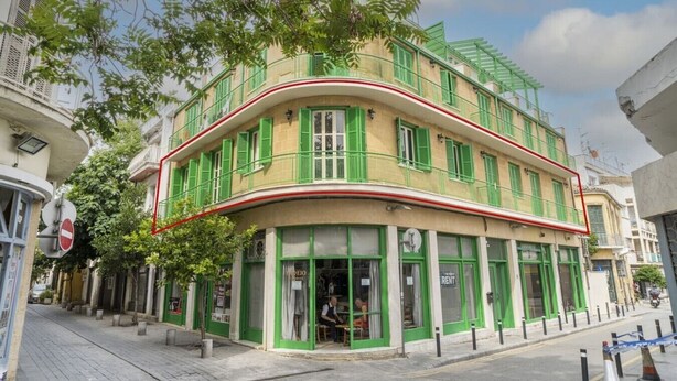 Un immeuble de deux étages doté d’une façade incurvée, de volets verts et d’un commerce situé au rez-de-chaussée, dont l’enseigne est bien visible.