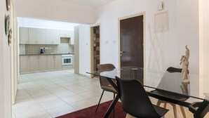 Une cuisine moderne avec des meubles blancs et un sol carrelé, un coin repas avec une table et des chaises en verre, et une porte sombre.