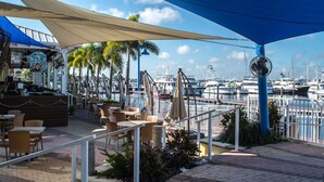 Un área de estar al aire libre con mesas y sillas, una sección cubierta con un letrero que dice 'Jolly Sailor Pub', y vistas de un puerto deportivo con barcos.