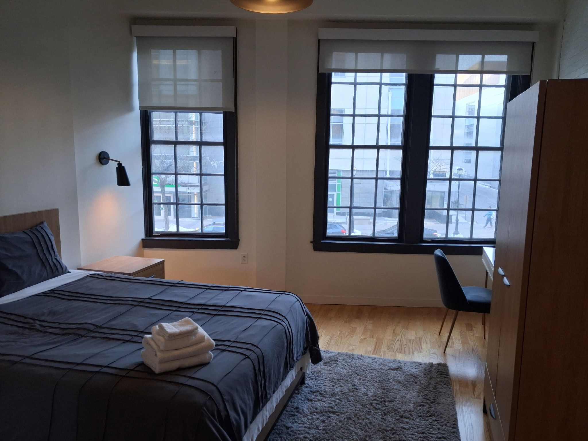 Quarto com cama, duas janelas, mesa de cabeceira, escrivaninha e guarda-roupa.