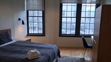 Quarto com cama, duas janelas, mesa de cabeceira, escrivaninha e guarda-roupa.