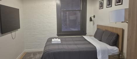 A bedroom with a bed, a window, a flat-screen TV, and a wall-mounted lamp.