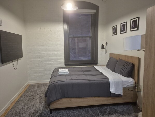 A bedroom with a bed, a window, a flat-screen TV, and a wall-mounted lamp.