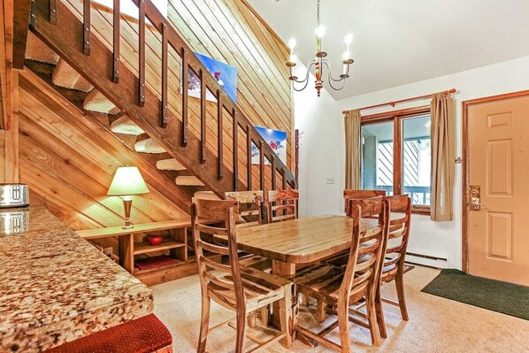 Un escalier en bois menant à une chambre aménagée en mezzanine, une table à manger avec des chaises, une lampe et une fenêtre avec des rideaux.