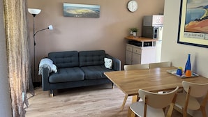 A living room with a sofa, a dining table with chairs, a lamp, a clock, and a framed picture on the wall.