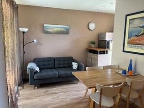 A living room with a sofa, a dining table with chairs, a lamp, a clock, and a framed picture on the wall.