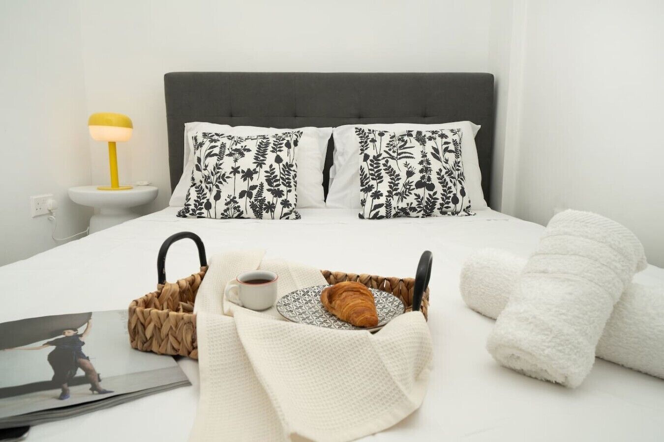 Ein ordentlich bezogenes Bett mit floralen Kissen, eine Nachttischlampe, ein Korb mit Croissants, eine Tasse Kaffee und gerollte Handtücher.