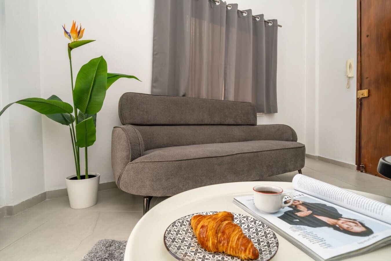 Una sala de estar con un sofá gris, una planta en maceta, una mesa con un croissant, una taza de café y una revista.