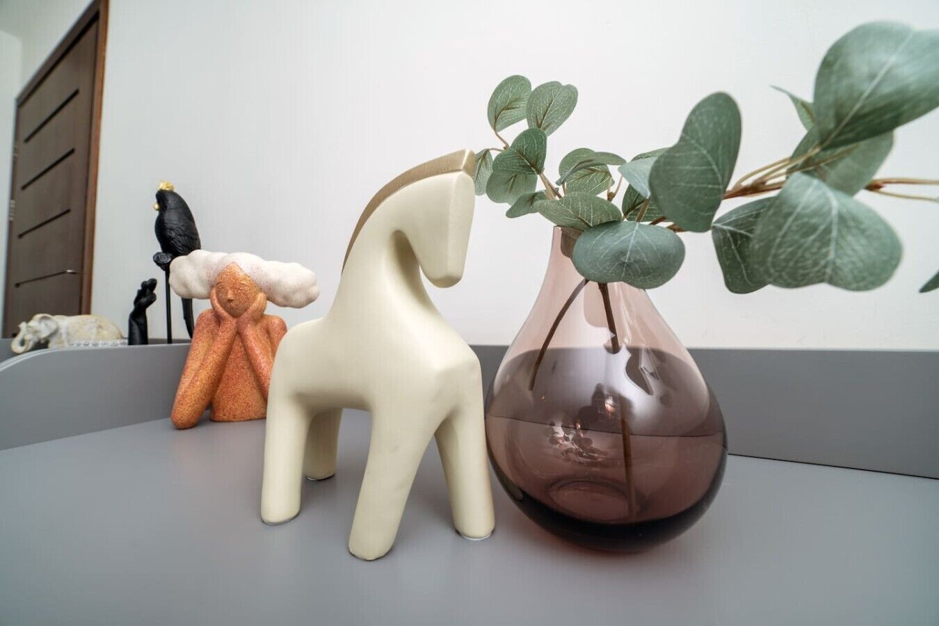A minimalist decor setup with a ceramic horse, a vase with green leaves, and a small sculpture on a grey surface.