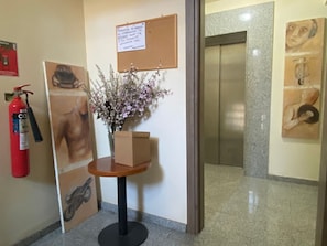 Zona de recepción con extintor, tablón de anuncios, un jarrón con flores, una mesa pequeña y un cartel en la pared.
