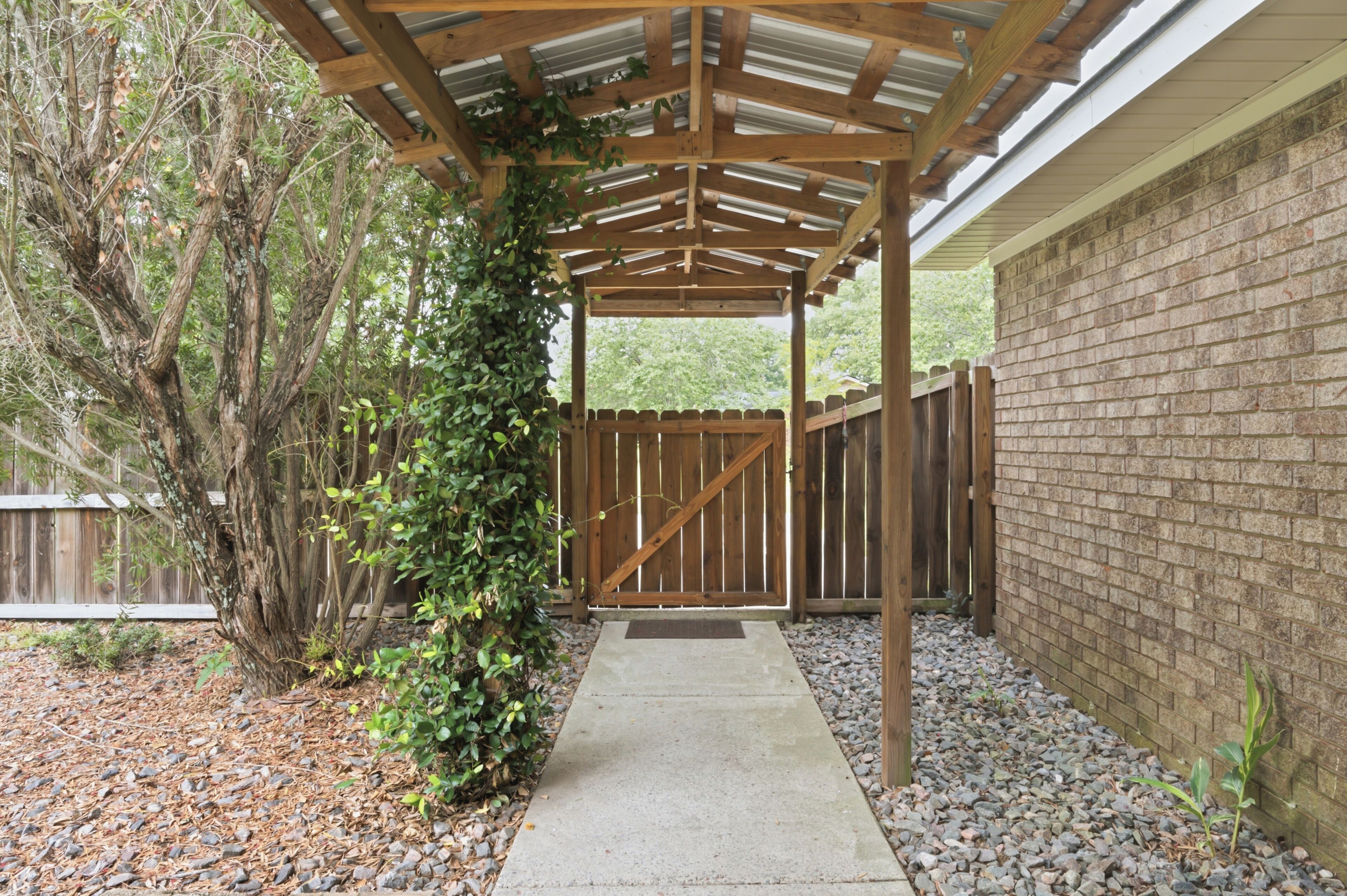 Un patio couvert, avec un chemin en béton menant à une porte en bois, le tout entouré d’un mur en briques et d’une clôture en bois.