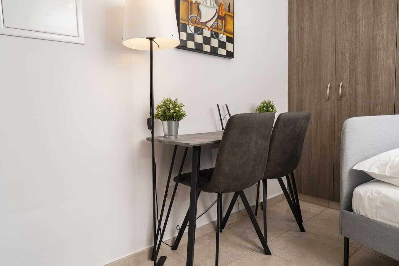 A small, modern room with a dining table, two chairs, a lamp, a bed, and a wardrobe.