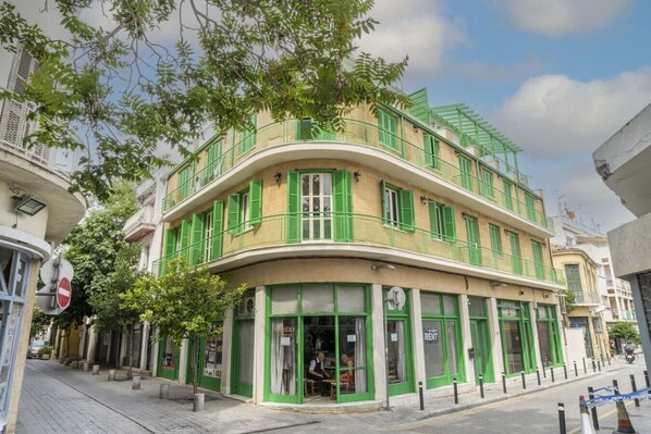 A multi-story building with green shutters, a curved facade, and a ground-level shop with a sign indicating it is for rent. - Room in shared flat in old town (Nicosia)