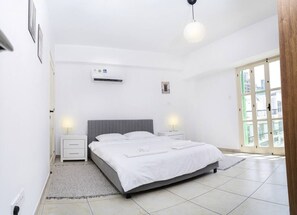 A bedroom with a bed, two bedside tables, an air conditioning unit, a window, and a door. - Room in shared flat in old town (Nicosia)