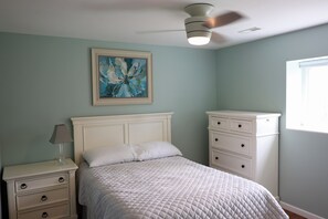 Ein Schlafzimmer mit Bett, Nachttisch, Schminktisch und Deckenventilator, sowie einem Bild an der Wand.