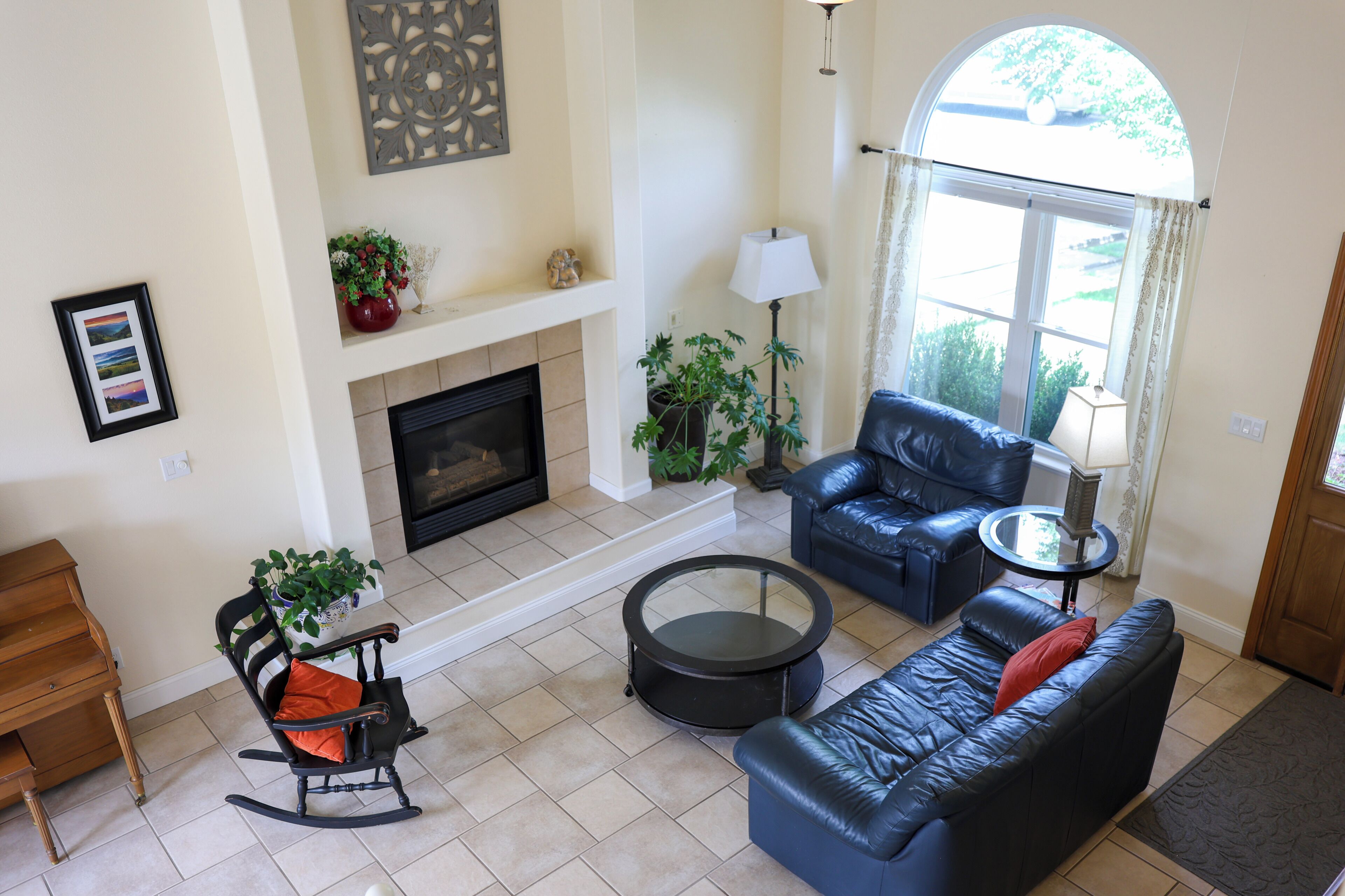 A living room with a fireplace, a black leather sofa, a rocking chair, a round coffee table, and a potted plant.