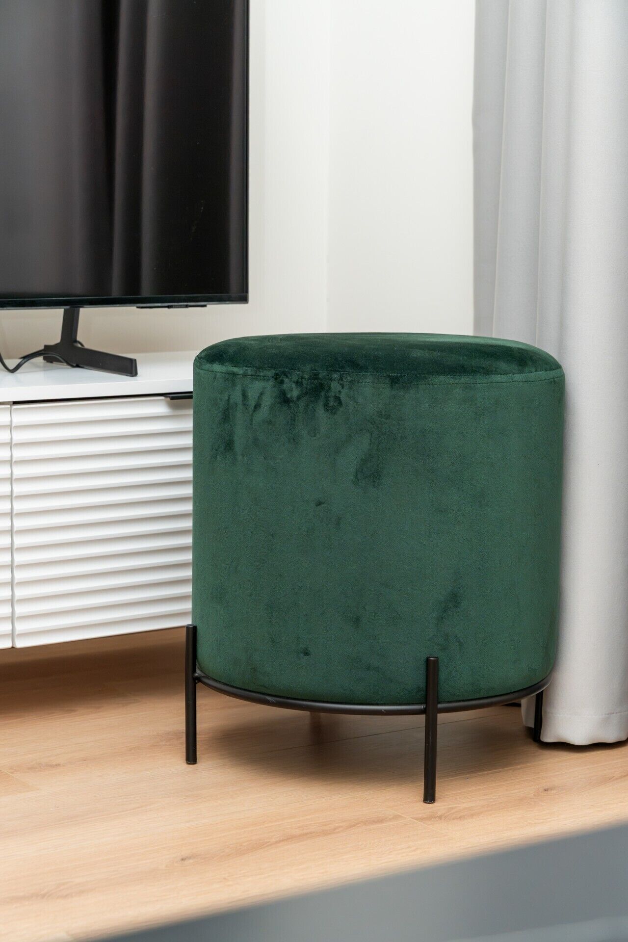 Un otomán verde de terciopelo con patas negras, ubicado frente a un armario blanco y un televisor.
