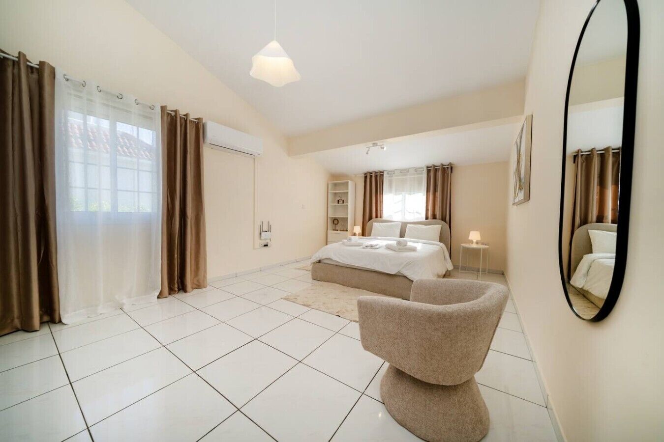 Quarto de hotel com cama, uma cadeira, um espelho e uma janela com cortinas.