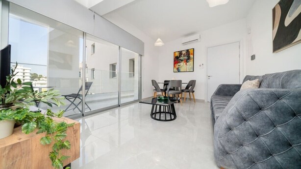 Een moderne woonkamer met een grijze bank, een eettafel met stoelen, een glazen scheidingswand en een balkon met planten.