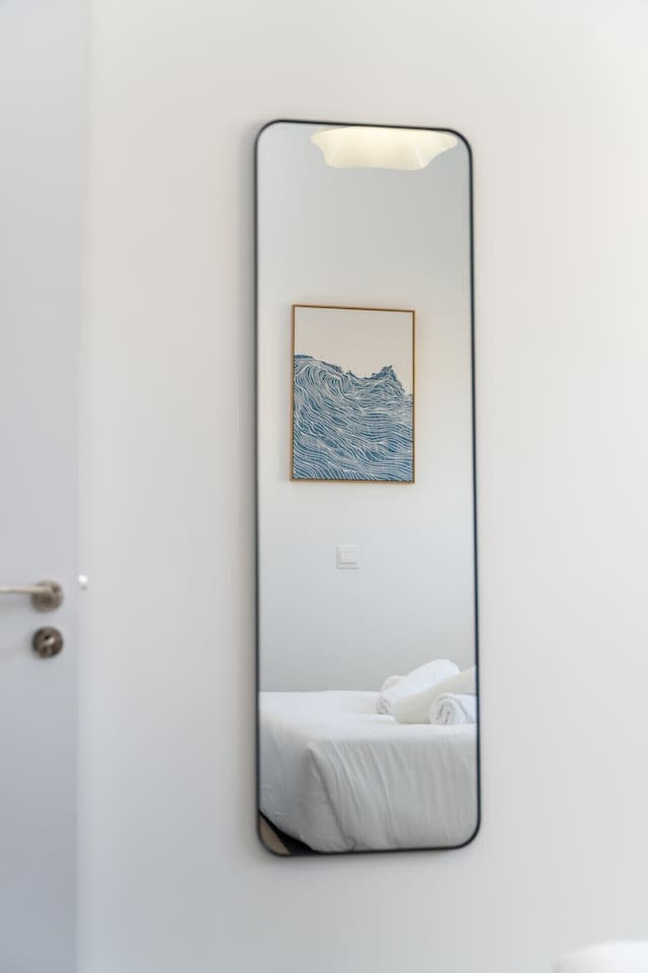 Un miroir orné d’une reproduction d’une œuvre d’art représentant un paysage de montagne, une porte blanche et un lit recouvert d’une couverture blanche.