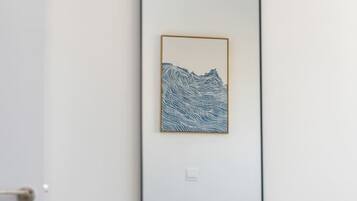 Un miroir orné d’une reproduction d’une œuvre d’art représentant un paysage de montagne, une porte blanche et un lit recouvert d’une couverture blanche.