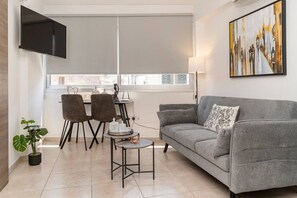 A living room with a grey sofa, a small table, two chairs, a TV mounted on the wall, and a painting. - Central 1 bedroom flat, near sea (Limassol)