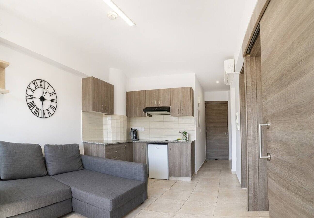 A modern living room with a grey sectional sofa, a kitchen with wooden cabinets, and a wall clock.