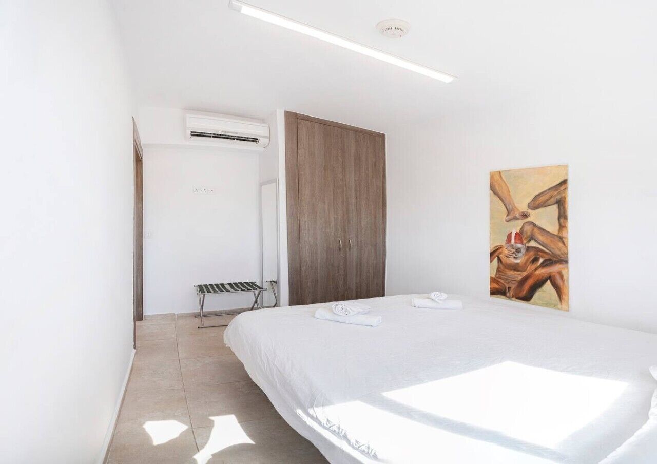 A bedroom with a bed, an air conditioning unit, a wooden wardrobe, a bench, and a painting on the wall.