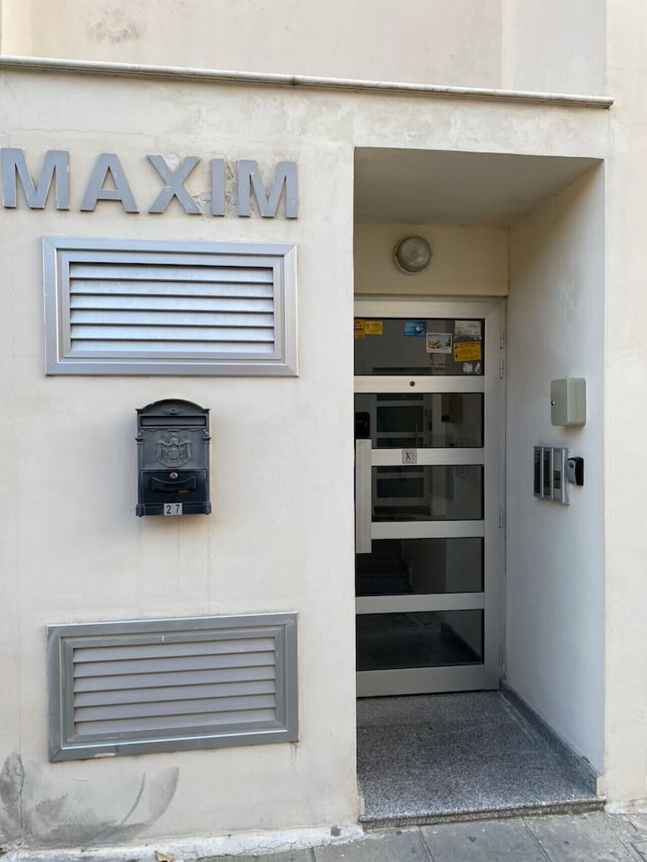 Entrada de un edificio con el nombre 'MAXIM' sobre un buzón y una puerta con un panel de vidrio.