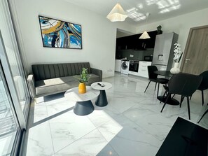 A modern living area with a sofa, two coffee tables, a dining table with chairs, a kitchen in the background, and a large abstract painting on the wall.