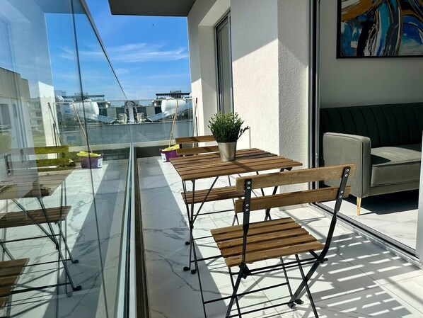 A balcony with wooden tables and chairs, a potted plant, and a view of buildings and a body of water.