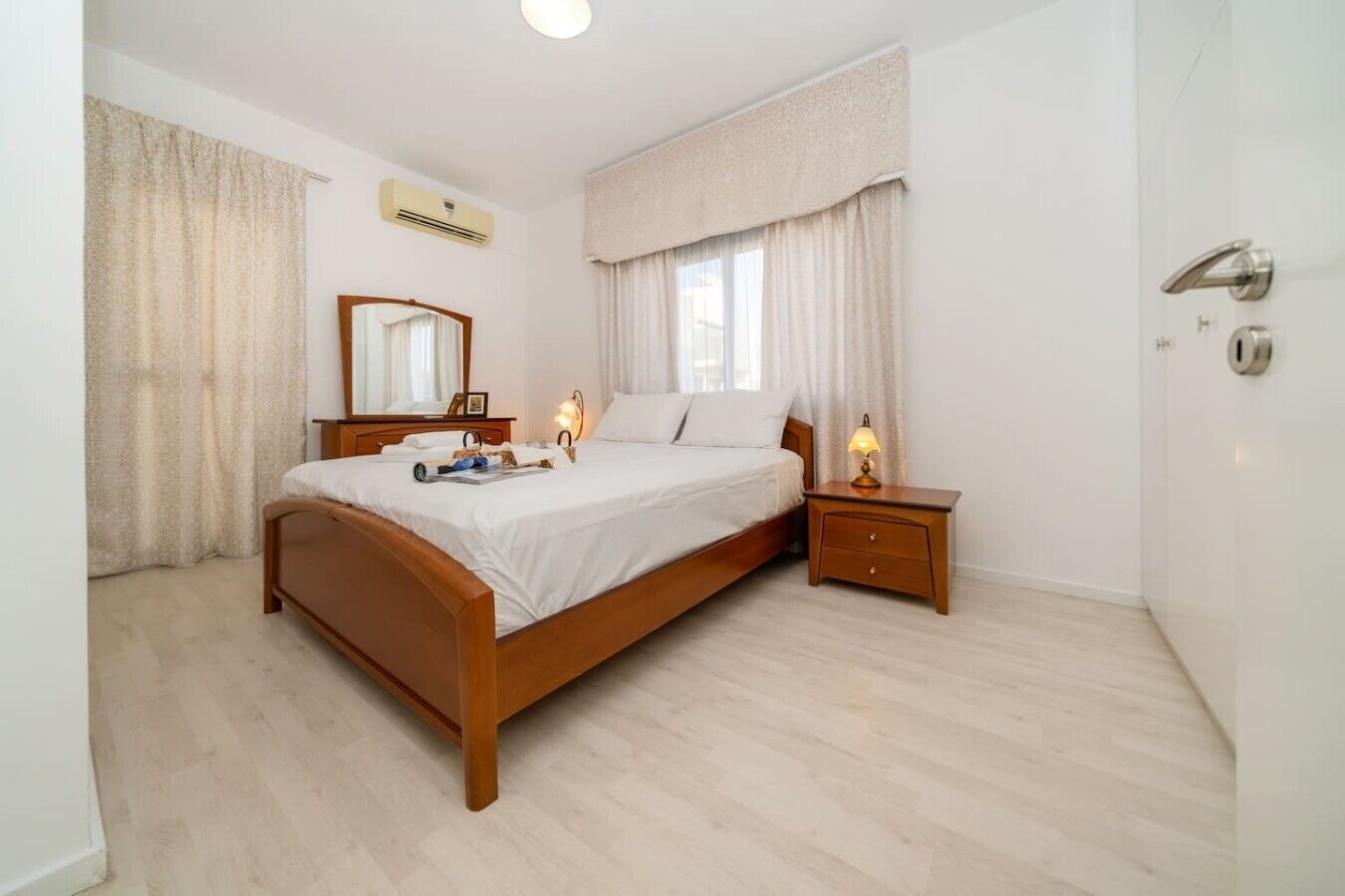 Une chambre à coucher avec un lit en bois, une table de chevet avec une lampe, un miroir et une fenêtre avec des rideaux.