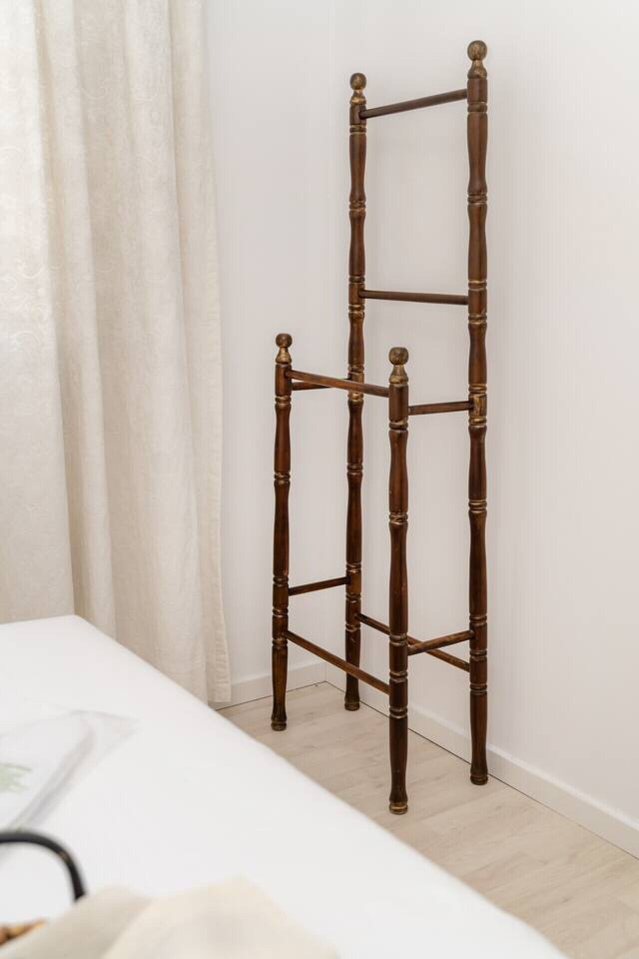 Ein hoher, freistehender Holzregal mit schlichtem Design, vor einer schlichten Wand in einem Zimmer mit einem weißen Bett und hellem Boden platziert.