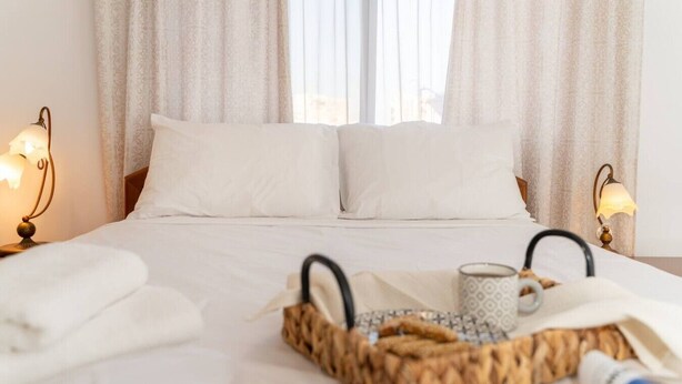 Ein Hotelzimmer mit einem Bett, zwei Kissen, einem Nachttisch mit Lampe, einem Tablett mit Handtüchern und einer Tasse sowie einem Fenster mit Vorhängen.