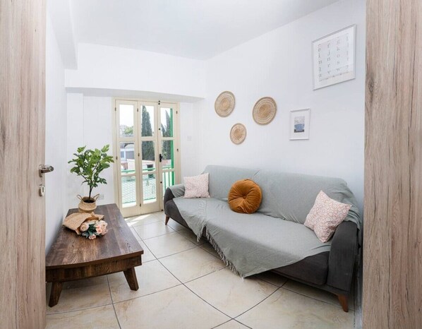 Ein Wohnzimmer mit grauem Sofa, einem Holztisch, einer Zimmerpflanze und einer Tür zu einem Außenbereich.