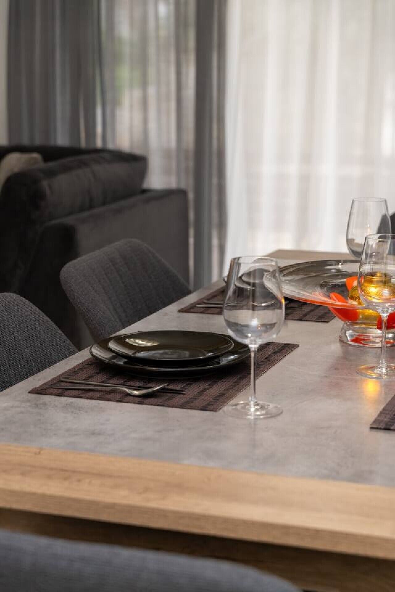 Een eettafel gedekt met borden, bestek en glazen, met een bank en gordijnen op de achtergrond.