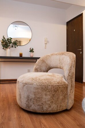 Un fauteuil rond, de couleur beige, avec un revêtement en tissu et un haut dossier, dans une pièce dont le sol est recouvert de planches de bois, avec un miroir rond accroché au mur et une porte en bois.