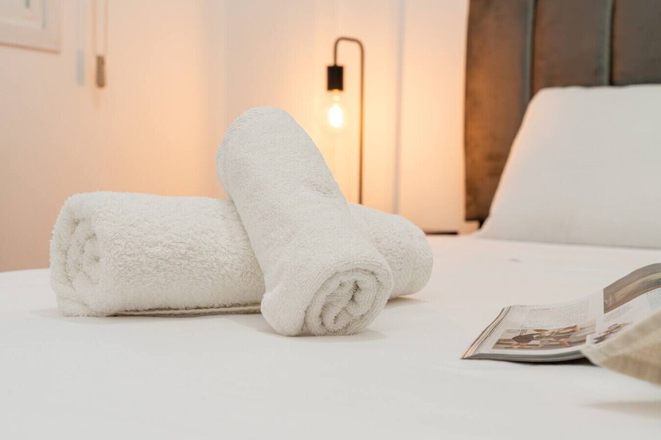 A neatly made bed with two rolled white towels and a magazine on it.