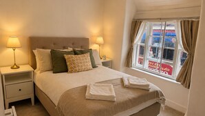 Una cama bien hecha con cubrecama y almohadas color beige, dos lámparas de noche, una ventana con cortinas y vista a la calle.