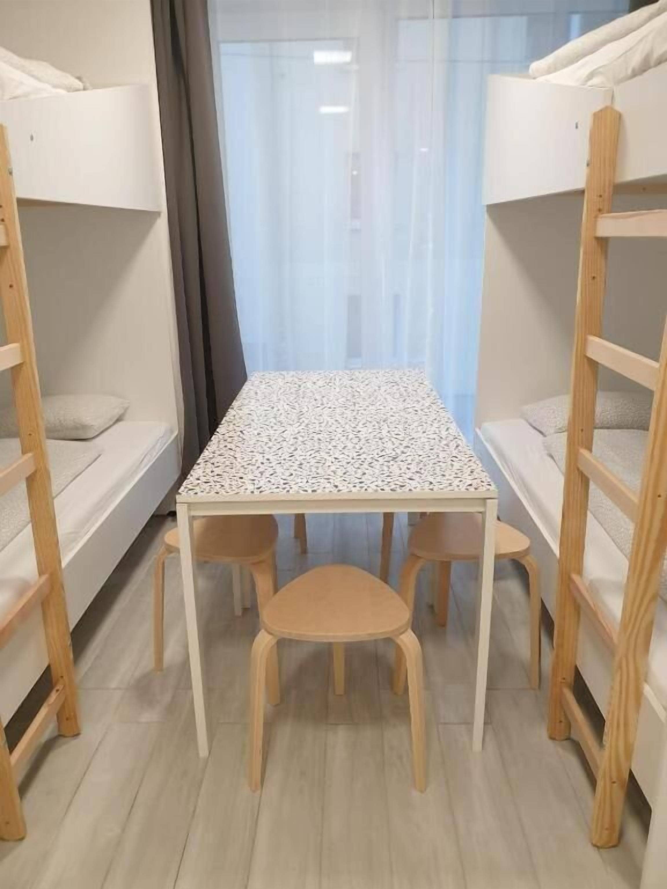 Ein Schlafraum mit Etagenbetten, einem kleinen Tisch und zwei Stühlen, einem Fenster mit Vorhängen und einem Holzboden.
