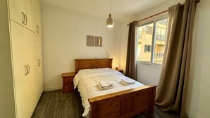 Une chambre à coucher avec un lit en bois, une table de chevet, une armoire, une fenêtre avec des rideaux et un luminaire suspendu.