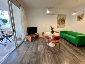 Een moderne woonkamer met een groene bank, een televisie, een eettafel met stoelen en een schuifdeur naar een buitenruimte.