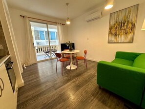 Een moderne woonkamer met een groene bank, een ronde eettafel met stoelen en een groot raam met gordijnen.